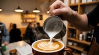 Barista pouring steamed milk creating latte art in coffee cup at cafe. Professional coffee making process with perfect foam technique for cappuccino preparation.