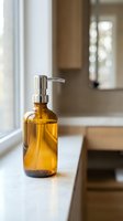 Amber glass soap dispenser with chrome pump on white marble countertop in modern kitchen interior with natural lighting and warm wood cabinetry.