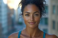 Young mixed race woman with curly hair in blue tank top smiling confidently at camera against blurred urban background, natural makeup and glowing skin.