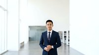Asian male professional in dark business suit and tie standing in modern office corridor with natural light from windows, suitable for corporate, business, and corporate culture concepts.