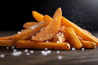 Golden french fries sprinkled with sea salt on dark wooden surface, falling salt crystals captured in motion, close-up food photography with dramatic lighting.