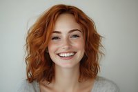 Young woman with natural red wavy hair and genuine smile wearing casual grey sweater against light background, radiating warmth and authenticity in studio portrait.