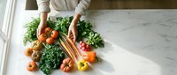 Overhead view of hands arranging fresh organic vegetables including carrots, kale, tomatoes and peppers on marble countertop for healthy cooking and meal preparation.
