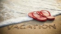 Red flip flops on sandy beach with vacation written in sand, ocean waves in background, summer holiday travel concept.