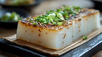 Seared Japanese turnip cake garnished with fresh green onions on bamboo mat. Close-up view shows caramelized top with crispy golden-brown crust and soft interior.