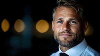 Confident mature Caucasian businessman with beard and styled blonde hair smiling at night, bokeh lights background creates professional atmosphere.