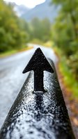 Black directional arrow on wet metal railing along mountain road, shallow depth of field with blurred forest background and rainy weather atmosphere.