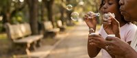 African American grandmother and granddaughter blowing soap bubbles together in sunny park, bonding across generations in outdoor family activity.