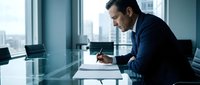 Professional businessman in dark suit writing documents at modern office desk with city skyline view through large windows for corporate presentations.