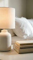 Warm glowing table lamp with cream ceramic base and linen shade beside stacked books on nightstand, cozy bedroom interior with white pillows and bedding in background for home decor and lifestyle.