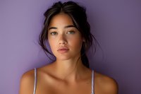 Young Asian woman with natural makeup and messy updo hairstyle looking at camera against purple background, wearing white tank top, beauty portrait with soft lighting.