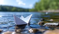 Paper boat floating on lake water surface with stones and bokeh light reflections, shallow depth of field nature background for childhood dreams and summer leisure.