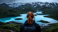 Young woman with backpack gazing at turquoise mountain lake surrounded by snow-capped peaks and green valleys, capturing wanderlust and outdoor adventure spirit.