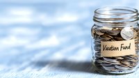 Glass jar filled with coins labeled vacation fund on blue background, representing savings goals and financial planning for travel and leisure activities.