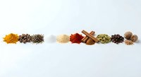 Colorful spices and seasonings arranged in neat piles on white background, including turmeric, peppercorns, cinnamon sticks and cardamom pods for cooking.
