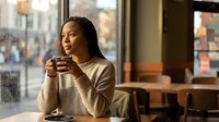Young African American woman enjoying hot coffee in cozy cafe by window on rainy day, peaceful morning moment with warm lighting and urban street view.