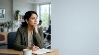 Young South Asian woman in olive green blazer and patterned blouse sitting at wooden desk, writing notes, thoughtful expression, modern office environment with plants and windows.