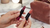 Woman hands holding two lipsticks in burgundy and coral shades near pink velvet cosmetic bag on marble surface for beauty and makeup routine.
