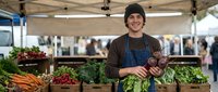 Young Hispanic male farmer vendor smiling at outdoor farmers market with fresh organic vegetables including carrots, radishes, leafy greens in wooden crates under tent.