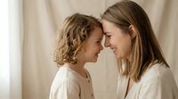 Tender moment between mother and daughter touching foreheads in soft natural light, expressing love and connection in cozy home setting.