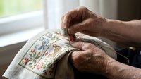 Elderly woman hands working on delicate floral embroidery hoop with colorful thread patterns. Traditional needlework craft hobby for relaxation and creativity.