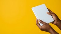 Hands of a person with dark skin tone holding a blank white book or notebook against a bright yellow background, suitable for publishing, education, and learning concepts.