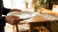 Close-up of hands holding empty white plate in warm home setting, suitable for food service, hospitality, dining, and restaurant marketing materials.