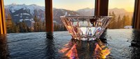 Crystal glass bowl on a dark wet surface with a stunning mountain sunset and snowy peaks visible through a large window in the background.