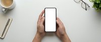 Male hands holding smartphone with blank white screen on clean white desk workspace with coffee cup, eyeglasses and plant for mockup design projects.