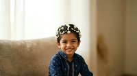 Young South Asian boy, approximately 3-5 years old, wearing a decorative floral crown and blue patterned clothing, smiling at camera while sitting on beige sofa.