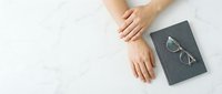 Hands resting on white marble surface next to closed dark notebook with eyeglasses, minimalist workspace setup for business planning and productivity.