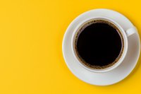 White ceramic cup filled with black coffee on bright yellow background, viewed from above. Minimalist composition with copy space for text or design elements.