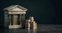 Miniature bank building with classical columns next to stacked coins on wooden surface representing financial services and monetary savings.