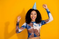 Excited young African American woman celebrating birthday with party hat and raised fists against bright orange background for festive occasions.