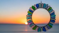 Colorful sea glass mirror with vibrant blue green purple pieces arranged in circular frame against golden sunset ocean horizon for coastal home decor.