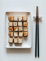 Pan-fried tofu cubes arranged in geometric pattern on white rectangular ceramic plate with black chopsticks, minimalist Asian cuisine presentation on light background.