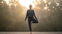 Young Caucasian woman practicing tree pose yoga outdoors in misty forest at sunrise, embodying balance, mindfulness and wellness.