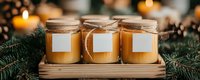 Handmade soy candles in glass jars with blank labels on wooden tray decorated with pine branches and glowing candlelight creating cozy atmosphere.