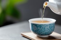 Pouring hot tea from white ceramic teapot into traditional blue patterned cup on wooden serving board against dark background with green bokeh effect.