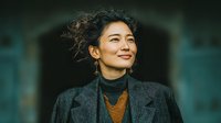 Professional Asian businesswoman in elegant dark suit smiling confidently with windswept hair against modern corporate building background for business success concepts.