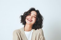 Young Asian woman with short wavy black hair laughing with genuine joy, wearing casual beige cardigan and white top against light background. Natural lifestyle portrait.