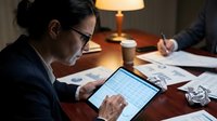 Professional businesswoman analyzing financial data on tablet during late evening work session with documents and coffee on wooden desk.
