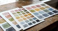 Watercolor paint palette sheets with vibrant color swatches spread on rustic wooden table for artistic projects and creative design work.