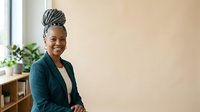 Young African American woman with braided updo hairstyle wearing dark teal blazer over cream top, smiling at camera in bright office with potted plants and wooden furniture.