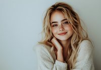 Young Caucasian woman with blonde wavy hair and natural makeup smiling warmly while wearing cozy white knit sweater against light background, expressing genuine happiness.