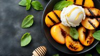 Grilled peach dessert with vanilla ice cream scoop and honey drizzle on black plate, garnished with fresh basil leaves against dark background. Food photography.