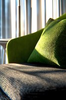 Sunlight streaming through window blinds creates dramatic shadows on green velvet armchair with textured cushion, capturing cozy home interior atmosphere.