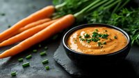 Fresh carrot cream soup garnished with chopped herbs in black bowl on dark stone background with whole carrots and greenery, healthy vegetarian meal.