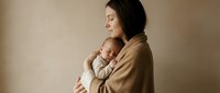 Young Caucasian mother in beige sweater tenderly holding sleeping newborn baby in warm embrace against neutral background for family portraits.