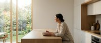 Young Asian woman sitting at modern kitchen island writing in notebook, peaceful morning routine in minimalist home with large windows overlooking garden.